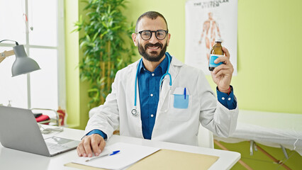Young hispanic man doctor using laptop holding medication bottle at clinic