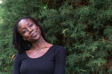 Young black woman with natural sister loc hairstyle smiling while standing in front of a pine tree at a park. No make up and wearing a black top