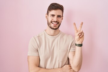 Hispanic man with beard standing over pink background smiling with happy face winking at the camera doing victory sign with fingers. number two.