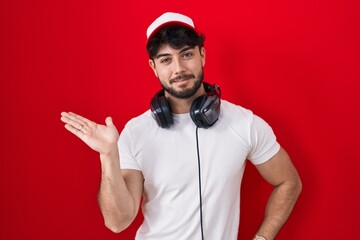 Hispanic man with beard wearing gamer hat and headphones smiling cheerful presenting and pointing with palm of hand looking at the camera. © Krakenimages.com