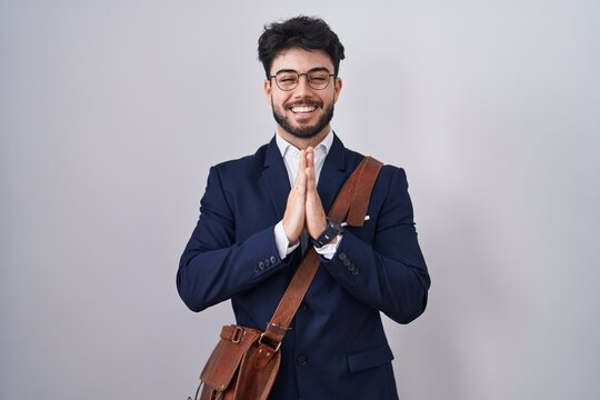 Hispanic man with beard wearing business clothes praying with hands together asking for forgiveness smiling confident.
