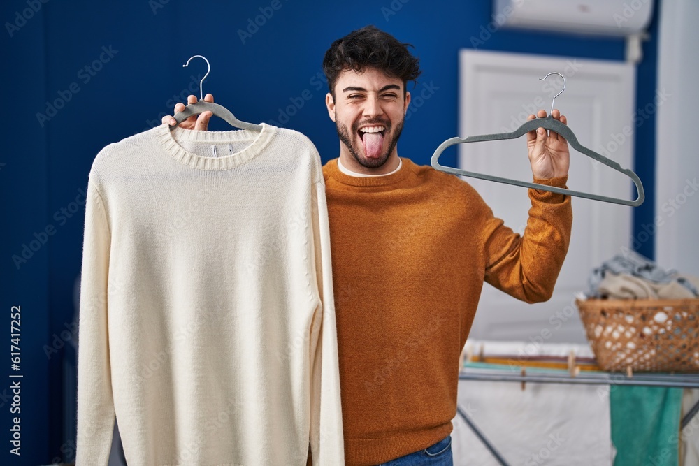 Sticker hispanic man with beard holding sweater on hanger at laundry room sticking tongue out happy with fun