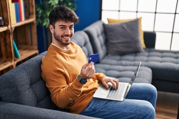 Young hispanic man using laptop and credit card sitting on sofa at home