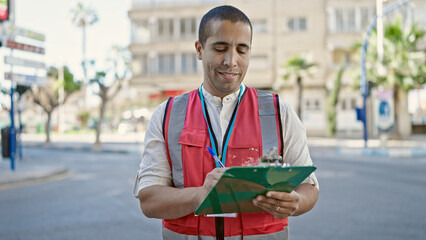 Young hispanic man volunteer smiling holding clipboard at street