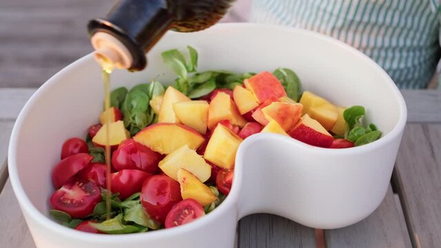 Close-up cooking summer salad during sunset on home terrace. The salad is drizzled with olive oil. Salad of cherry tomatoes, strawberries, herbs, peach in a large white designer bowl