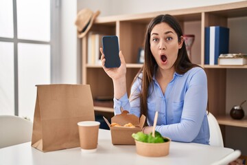 Young brunette woman eating take away food at home showing smartphone screen scared and amazed with...