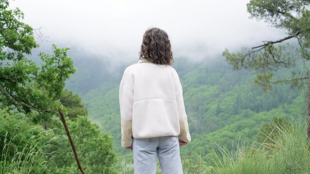 Portrait young woman in fleece jacket and jeans stands on top of a hill overlooking mountains trees clouds. Female person stretching and admires the picturesque view.  Video from the back
