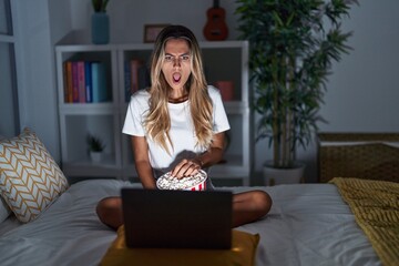 Young blonde woman sitting on the bed at home watching a movie from laptop in shock face, looking skeptical and sarcastic, surprised with open mouth
