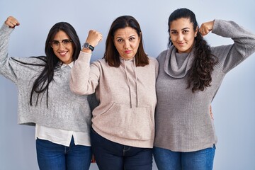 Mother and two daughters standing over blue background strong person showing arm muscle, confident and proud of power