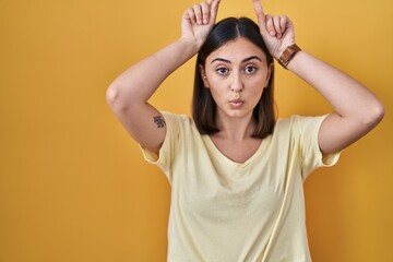 Fototapeta na wymiar Hispanic girl wearing casual t shirt over yellow background doing funny gesture with finger over head as bull horns