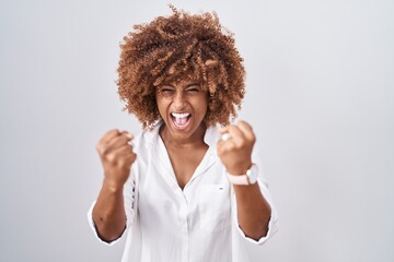 Young hispanic woman with curly hair standing over white background angry and mad raising fists...
