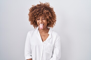 Young hispanic woman with curly hair standing over white background sticking tongue out happy with...