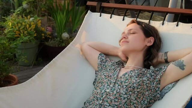 Portrait of young carefree woman enjoying a sunny day in the hammock on her terrace. Female resting stretching after work and enjoying beautiful weather sunset