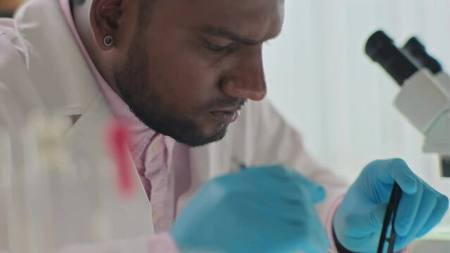 Close up of microbiologist in protective uniform using tweezers and petri dish working at research in laboratory