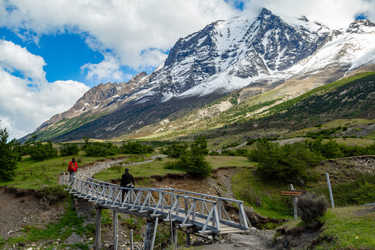 Paisagem das montanhas nevadas dos Andes no Chile com dois turista fazendo trilha, atravessando a ponte de madeira abaixo em dia ensolarado de c&eacute;u zul com nuvens.