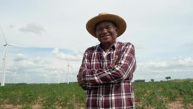 Portrait of elderly male farmer smiling and crossed arms while standing in field, outdoors, with wind farm in background.
