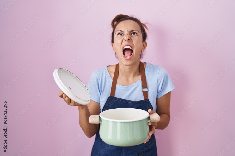 Poster brunette woman wearing apron holding cooking pot angry and mad screaming frustrated and furious, sho