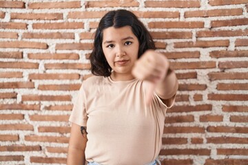 Young hispanic woman standing over bricks wall looking unhappy and angry showing rejection and negative with thumbs down gesture. bad expression.