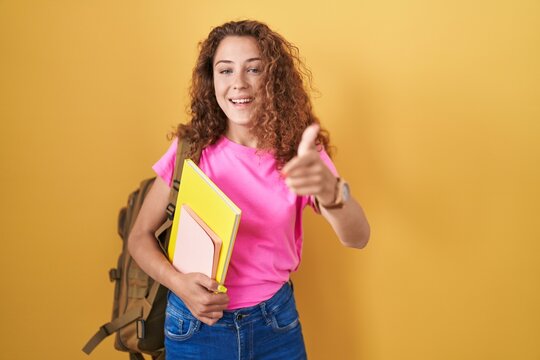 Young caucasian woman wearing student backpack and holding books pointing fingers to camera with happy and funny face. good energy and vibes.