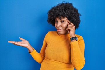 Fototapeta na wymiar Black woman with curly hair standing over blue background confused and annoyed with open palm showing copy space and pointing finger to forehead. think about it.