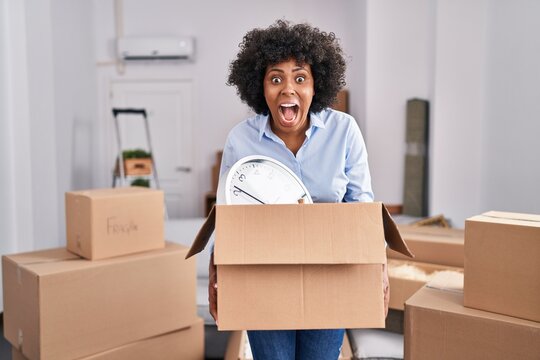 Black woman with curly hair moving to a new home holding cardboard box angry and mad screaming frustrated and furious, shouting with anger looking up.