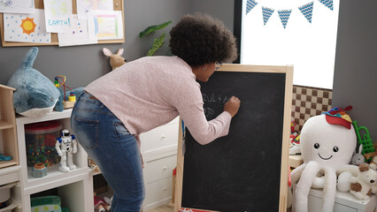 Young african american woman preschool teacher writing on blackboard at kindergarten
