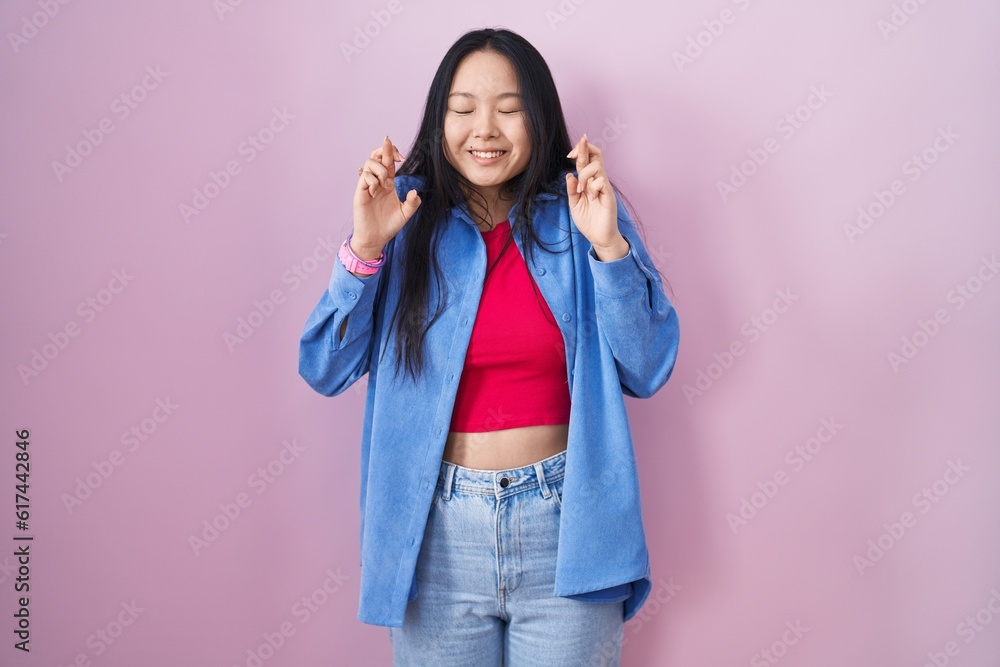 Poster Young asian woman standing over pink background gesturing finger crossed smiling with hope and eyes closed. luck and superstitious concept.