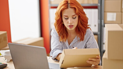 Young redhead woman ecommerce business worker using laptop writing on package at office