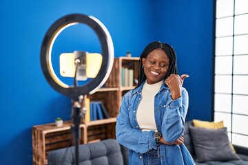 African woman with braids recording vlog tutorial with smartphone at home pointing thumb up to the...