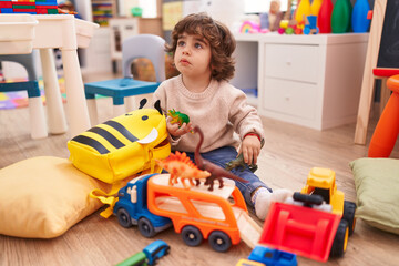Adorable hispanic boy playing with car and dinosaur toy sitting on floor at kindergarten