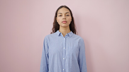 Young beautiful hispanic woman standing with serious expression over isolated pink background