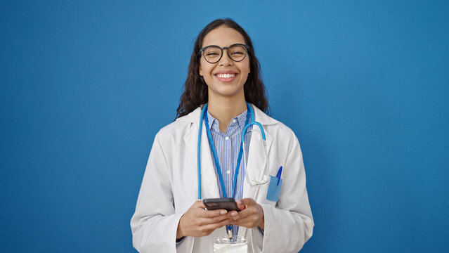 Young beautiful hispanic woman doctor smiling using smartphone over isolated blue background