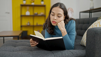 Young beautiful hispanic woman reading book lying on sofa at home