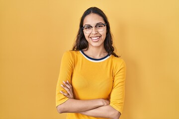Obraz premium Young brazilian woman wearing glasses over yellow background happy face smiling with crossed arms looking at the camera. positive person.