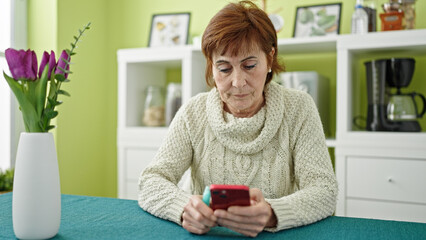 Mature hispanic woman holding vape inhaler sitting on table using smartphone at dinning room