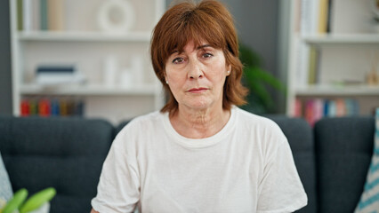 Middle age woman sitting on sofa with serious face home - obrazy, fototapety, plakaty