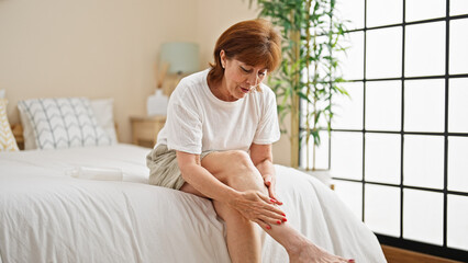 Middle age woman applying lotion on leg sitting on bed at bedroom