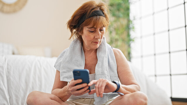 Middle age woman sitting on yoga mat using smartphone and watch at bedroom - Powered by Adobe