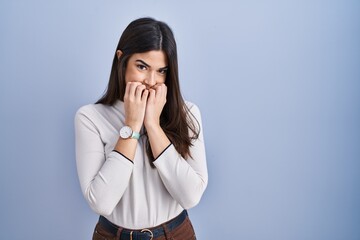 Young brunette woman standing over blue background laughing and embarrassed giggle covering mouth...