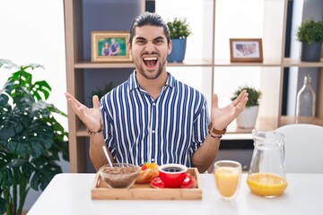 Fototapeta na wymiar Hispanic man with long hair sitting on the table having breakfast crazy and mad shouting and yelling with aggressive expression and arms raised. frustration concept.