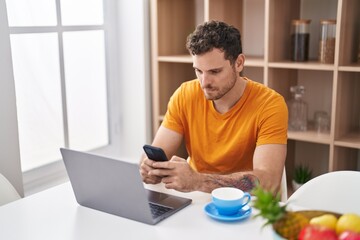 Young hispanic man using laptop and smartphone sitting on table at home