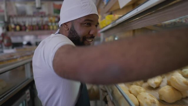 One black Brazilian employee working at food deli grabbing bread behind counter at bakery