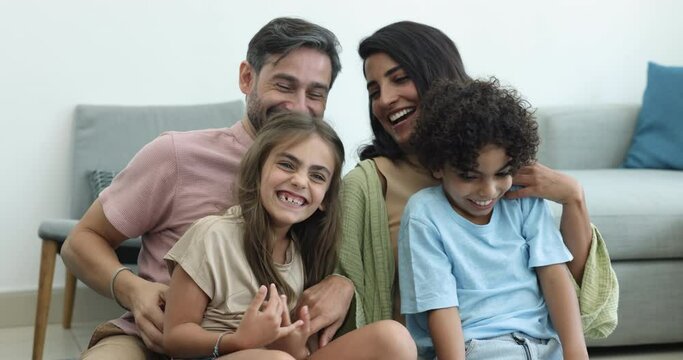 Happy married multinational couple pose for photo with cute siblings, tickling, laughing having fun sit on couch. Affectionate bonding multiethnic family enjoy leisure time at home. Playtime, custody