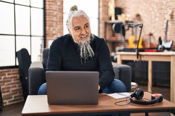 Middle age grey-haired man musician using laptop sitting on chair at music studio