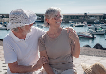 Fototapeta premium Smiling bonding senior family couple sitting together on bench close to the sea. Elderly man and woman hugging cuddling. Harbor and boats on background
