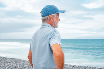 Rear view of senior man in casual polo shirt standing at the beach looking at horizon over sea,...