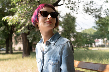 Woman with pink hair sitting on a park bench with sunglasses in summer