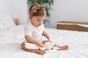 Adorable caucasian baby sitting on bed playing with doll at bedroom