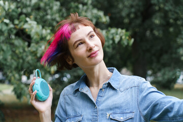 A girl with pink hair listens to music through the portable musical speaker in the park in summer