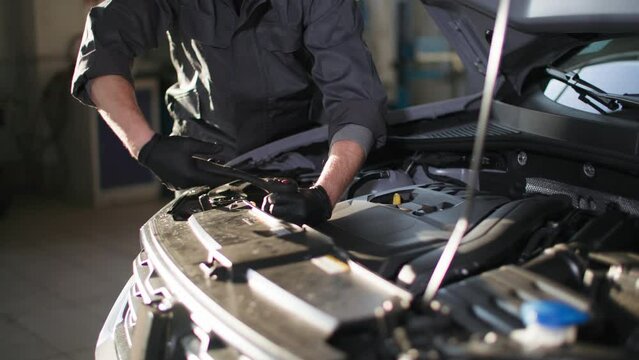 automobile service, young mechanic with wrench checks car engine near open hood in a car service
